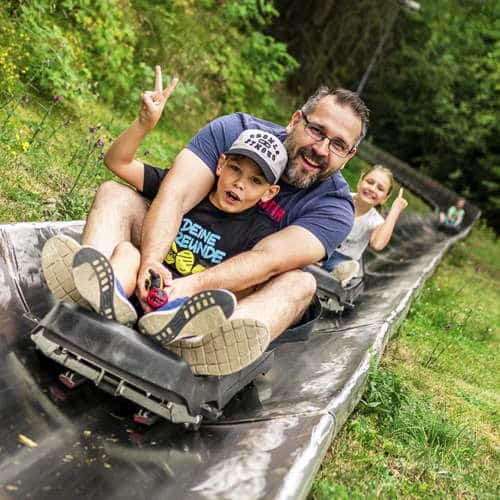 Ein Vater mit Bart und ein Junge flitzen auf einer Sommerrodelbahn mit sichtlich viel Spaß den Hang hinab.